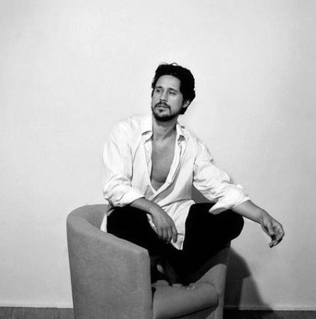 A black and white picture of Peter Gadiot sitting alone in a room.